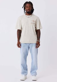 T-shirt oversize beige chiaro con una texture sbiadita, caratterizzata da una stampa logo marrone. Abbinato a jeans larghi blu chiaro e sneaker bianche.