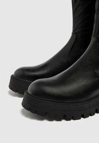 Bottes en cuir noir avec bout arrondi, dotées de semelles épaisses en caoutchouc noir texturé avec des crampons profonds, conçues pour la durabilité et l'adhérence.