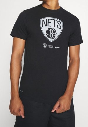 Person, die ein schwarzes T-Shirt mit dem Brooklyn Nets Basketball-Logo und schwarze Shorts trägt, steht mit den Händen nahe den Taschen vor einem einfarbigen Hintergrund.