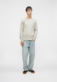 Jovem de pé, vestindo uma camisa de mangas longas bege, jeans azul claro e ténis pretos com atacadores brancos, contra um fundo cinza liso.