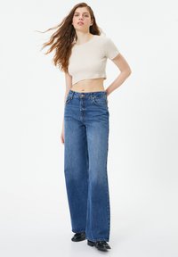 Korte lichtbeige top gecombineerd met hoge taille, wijde blauwe denim jeans. De jeans hebben klassieke zakken en een knoopsluiting.