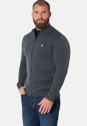 Man with beard wearing a dark gray zip-up sweater and blue jeans, standing with one hand in pocket against a plain background.