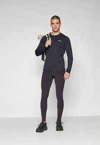 Zwart sportshirt met lange mouwen en logo, gecombineerd met zwarte leggings en zwarte sneakers. Model draagt een lichtgekleurde rugzak.