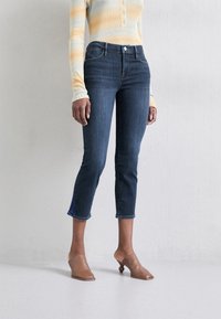 Jeans de mezclilla azul oscuro con corte ajustado en los tobillos, cintura alta y detalles de costura sutiles. Combinados con una blusa de rayas pastel y zuecos marrones.