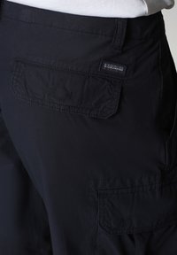 Shorts cargo noirs avec poche à rabat arrière et petite étiquette de marque sur la ceinture, portés avec une chemise blanche.