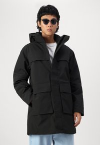 Black waterproof parka with a high collar, front zipper, and two large chest pockets. Worn over a white shirt, paired with light blue jeans.