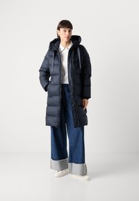 Mørkeblå polstret frakk med hette, med full glidelås og flere lommer, kombinert med vide jeans og lyse joggesko.