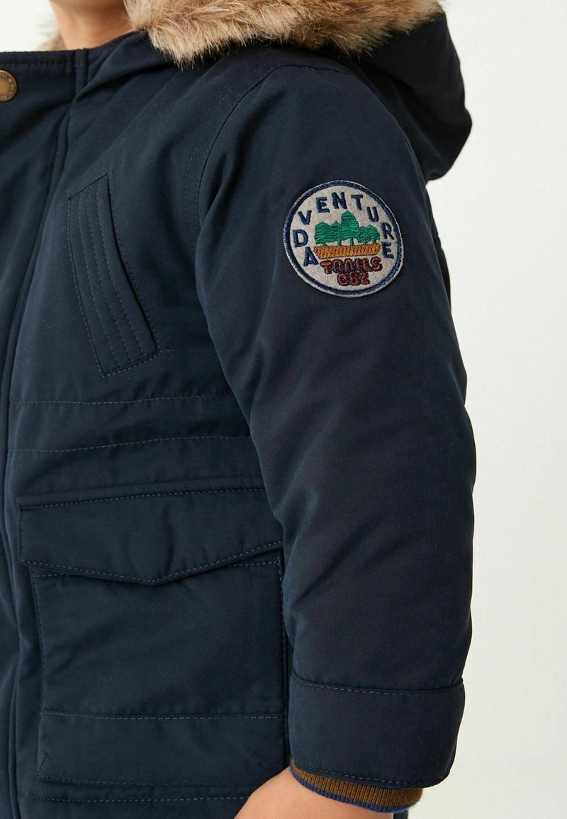 Enfant portant une veste d'hiver bleu foncé avec une capuche bordée de fourrure et un patch rond « Adventure Trails 082 » sur la manche.