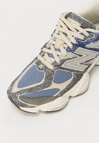 Blå och grå atletisk sko med ett mesh-övre, mockadetaljer, texturerad detaljering och krämfärgade snören. Gummisula för bra grepp.