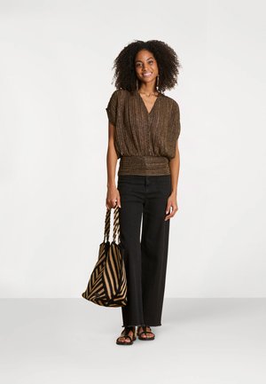 Femme souriante aux cheveux bouclés portant un haut bronze scintillant, un pantalon large noir, des sandales noires et tenant un grand sac fourre-tout rayé.
