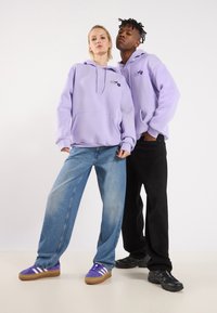 Lyse lilla hettegensere med frontlommer, brukt sammen med blå denim og svarte bukser. Distinkt blomsterbroderi på hettegensere, paret med sneakers.