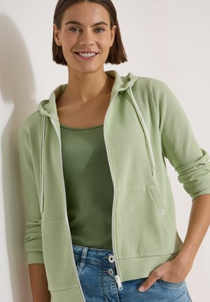 Femme souriante portant un sweat à capuche zippé vert clair sur un débardeur vert avec un jean bleu, main dans la poche, sur un fond uni.