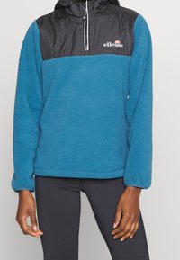 Pullover in pile blu con una parte superiore in nylon nero, zip metà lunghezza sul davanti e il logo "ellesse" stampato sul petto. Le maniche lunghe terminano ai polsi.