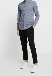 Uomo che indossa una camicia a maniche lunghe slim-fit con motivo, pantaloni neri a sigaretta e sneakers bianche con velcro, in piedi con una mano in tasca.