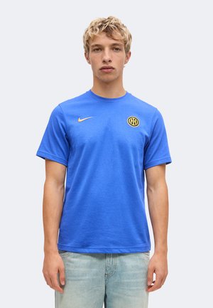 Mladý muž so svetlými kučeravými vlasmi, oblečený v modré tričko Nike so žltým logom Inter Miláno a svetlomodrých džínsach, stojaci otočený tvárou vpred.