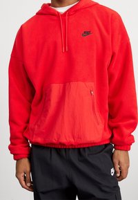 Felpa rossa in poliestere con tasca a marsupio e tasca laterale con zip. Cappuccio con cordino e orlo elasticizzato. Presenta un logo Nike nero sul petto.