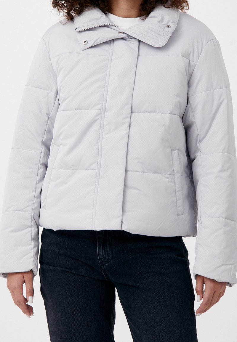 Femme portant une doudoune matelassée gris clair avec des poches latérales et un col montant zippé, associée à un pantalon noir.