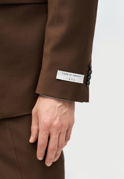 Χέρι με μανίκι καφέ σακακιού με ετικέτα "Tiger of Sweden 1903" και τέσσερα μαύρα κουμπιά στο μανίκι, πάνω σε φωτεινό φόντο.