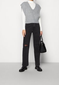 Grå stickad ärmlös topp över en vit långärmad skjorta, tillsammans med svarta slitna jeans och svarta ankelstövlar. Svart handväska ingår.