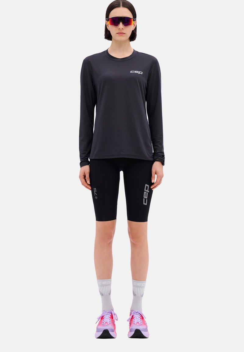 Schwarzes langärmeliges Sporthemd, schwarze Kompressionsshorts, graue Socken, pink- und grau-farbene Laufschuhe. CEP-Logo auf Hemd und Shorts.