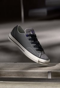 Grå sneaker med et glatt yttermateriale, suede tåhette, svarte snører, hvit gummisåle og lilla merkevaredetalj på tungen.