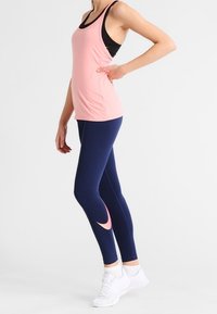 Kvinna står med en hand på höften, klädd i en rosa linne, marinblå leggings med en rosa swoosh, vita strumpor och vita sneakers.