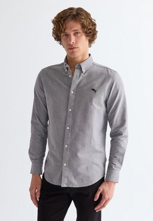 Junger Mann mit lockigem Haar, der ein hellgraues, geknöpftes Langarmhemd und schwarze Hose vor einem schlichten weißen Hintergrund trägt.