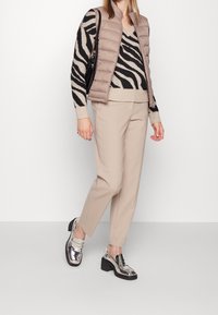 Texturovaná béžová péřová vesta přes černo-béžový svetr se zebřím vzorem, doplněná o světle béžové kalhoty a lesklé stříbrné mokasíny.