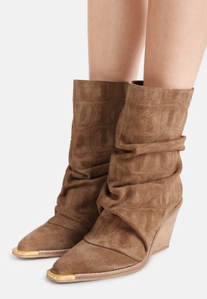 Braune Wildleder-Stiefel in lässiger Mid-Calf-Länge mit spitzen Zehen, goldfarbenen Metallkappen an den Zehen und gestapelten Keilabsätzen an den Beinen einer Person.