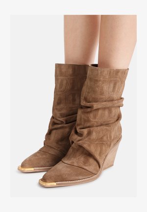 Stivali marroni in camoscio morbidi fino al polpaccio con punte appuntite, copritacchi in metallo dorato e tacchi a zeppa impilati indossati sulle gambe di una persona.