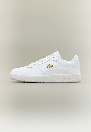 Fehér bőr sportcipők alacsony fazonnal, kerek orral, fűzős záródással és arany Lacoste logóval az oldalon.