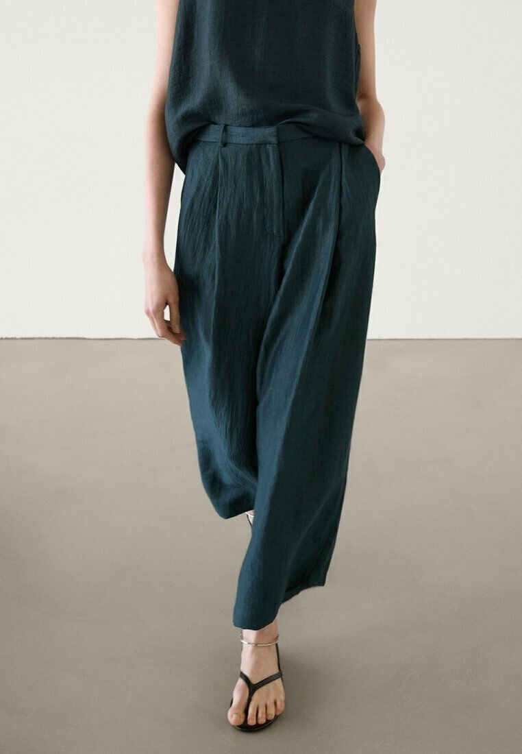 Dark teal, loose-fitting pants made of lightweight fabric, featuring a straight cut, pleats, and side pockets. Worn with a sleeveless top.