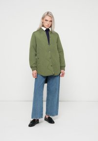 Olīvu zaļš quilted jaka ar pogu priekšpusi, sānu kabatām un brīvu piegriezumu, apvienota ar platām zilām džinsām un melnām kurpēm.