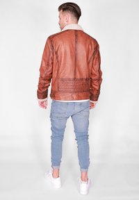 Bruine leren jas met een schapenvachtkraag, gestructureerde afwerking en stikdetails, gedragen over lichtblauwe jeans en witte sneakers.