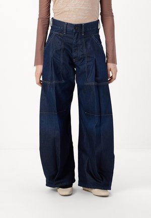 Jeansy z ciemnoniebieskiego denimu o szerokich nogawkach z przednimi kieszeniami naszywanymi, noszone z beżowymi butami i dopasowanym brązowym topem z długim rękawem.