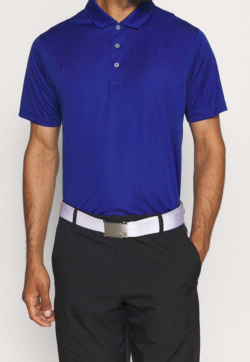 Polo bleu à manches courtes avec col et patte à trois boutons, porté avec un pantalon noir et une ceinture blanche avec une boucle argentée.