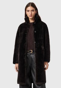 Casaco escuro de pelo sintético com gola, fecho de botões à frente e bolsos laterais. Combinado com um suéter castanho e calças de couro pretas.