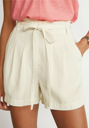 Femme portant un short blanc taille haute avec une ceinture en tissu nouée et un haut rose, une main dans la poche, montrant une montre-bracelet et un bracelet.