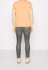 Camiseta de manga corta en color naranja claro, combinada con jeans gris ajustados y zapatillas blancas con suela de goma, vista desde atrás.