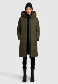 Olivovo zelený, pod kolená dlhý zateplený kabát s kapucňou, so zipsovým uzáverom a bočnými vreckami, skombinovaný s čiernymi legínami a členkovými čižmami.
