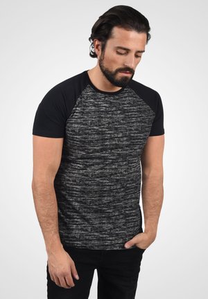 Camiseta de manga corta en gris y negro melange con escote redondeado y mangas raglán negras. Combinada con pantalones negros, el modelo posa con la cabeza ligeramente inclinada hacia abajo.