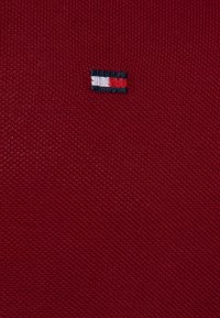 Červená pletená látka s malým vyšitým logem v námořnické modré, bílé a červené pruhy. Textura je strukturovaná a těsně tkaná.