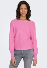 Maglione rosa a coste con scollo ampio e maniche lunghe, abbinato a jeans grigi. Il tessuto sembra morbido con una finitura testurizzata.