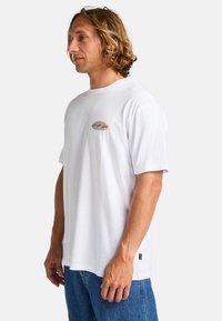 Hvid bomulds t-shirt med korte ærmer, som har et lille, farverigt bølgelogo på brystet. Båret med blå denim jeans.
