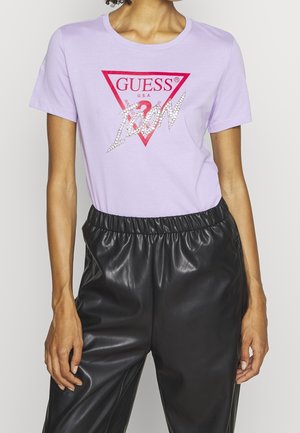 Woman wearing a lavender Guess t-shirt with red triangle logo and rhinestone "Icon" text, paired with black faux leather pants.