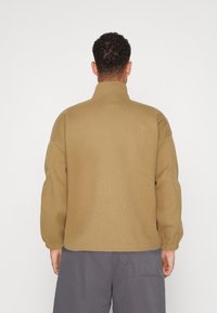 Brun tekstureret pullover med høj krave og elastiske manchetter, båret over grå bukser, med et enkelt og afslappet design.