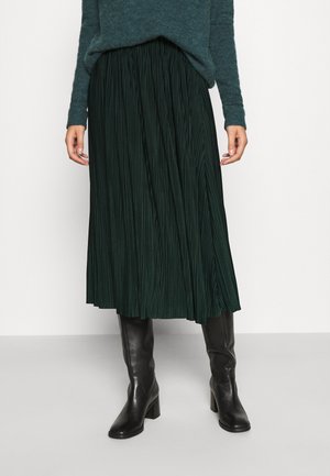 Femme portant une jupe midi plissée vert foncé, un pull bleu sarcelle et des bottes en cuir noires montantes, debout devant un fond uni.