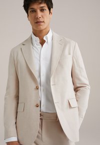Lys beige linned jakkesæt med to træknapper, notch lapeller og en brystlomme. Båret over en hvid skjorte med knapper.