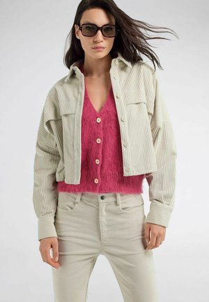 Donna che indossa una giacca e pantaloni in velluto a coste beige oversize con un gilet rosa acceso peloso abbottonato e occhiali da sole neri.