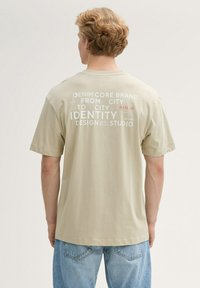 Beige katoenen T-shirt met korte mouwen, met witte tekst op de achterkant: branding en ontwerpdetails in verschillende lettertypen. Spijkerbroek zichtbaar.